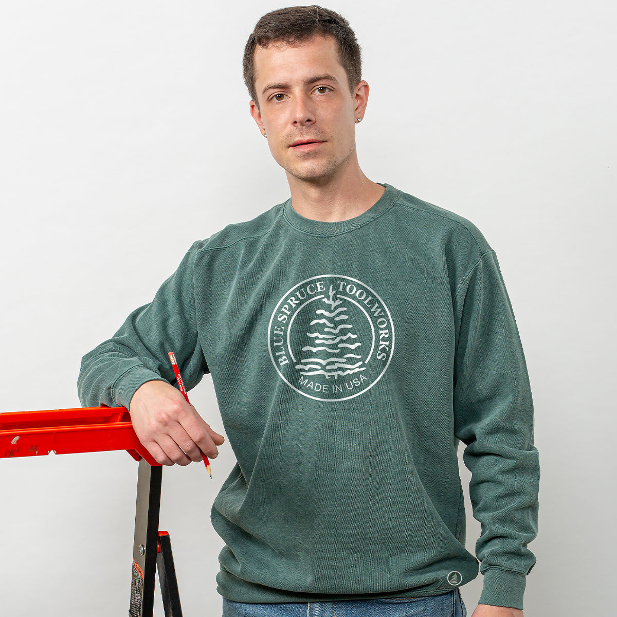 A man wearing the Blue Spruce crewneck sweatshirt holding a pencil and leaning against a ladder.