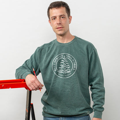 A man wearing the Blue Spruce crewneck sweatshirt holding a pencil and leaning against a ladder. 