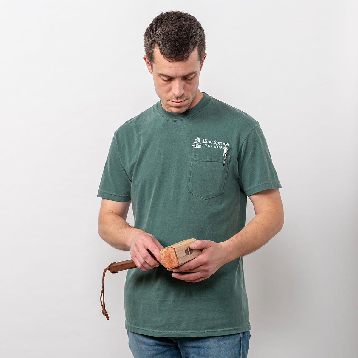 A man standing, holding a mallet with the front of the Blue Spruce T-shirt facing the camera. 